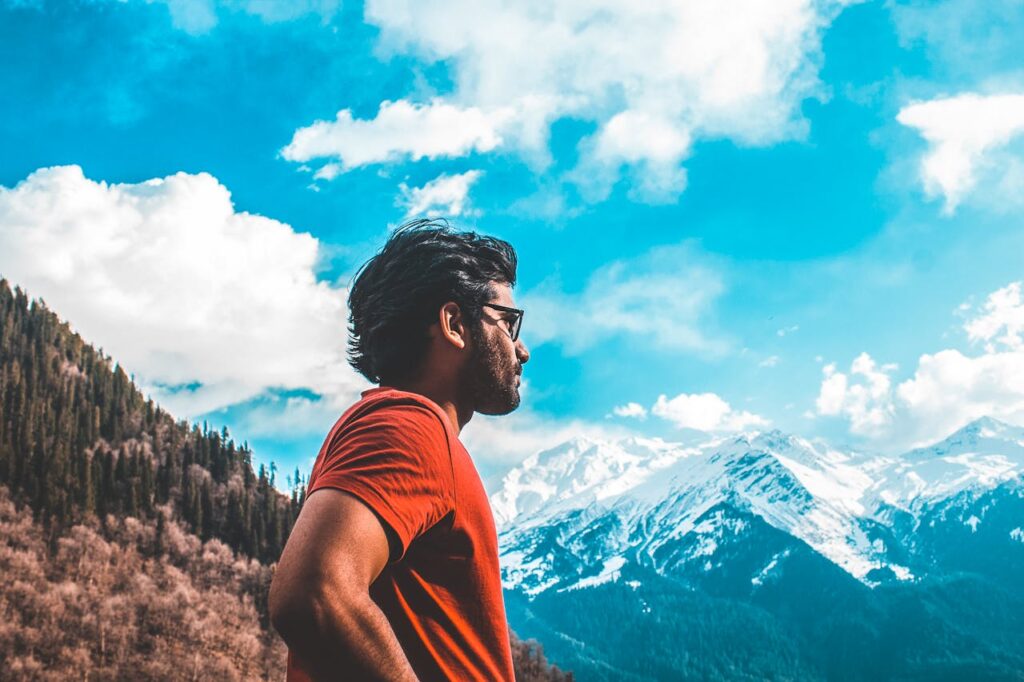 A man in a red shirt stands outdoors, enjoying a scenic mountain view under a vibrant blue sky.